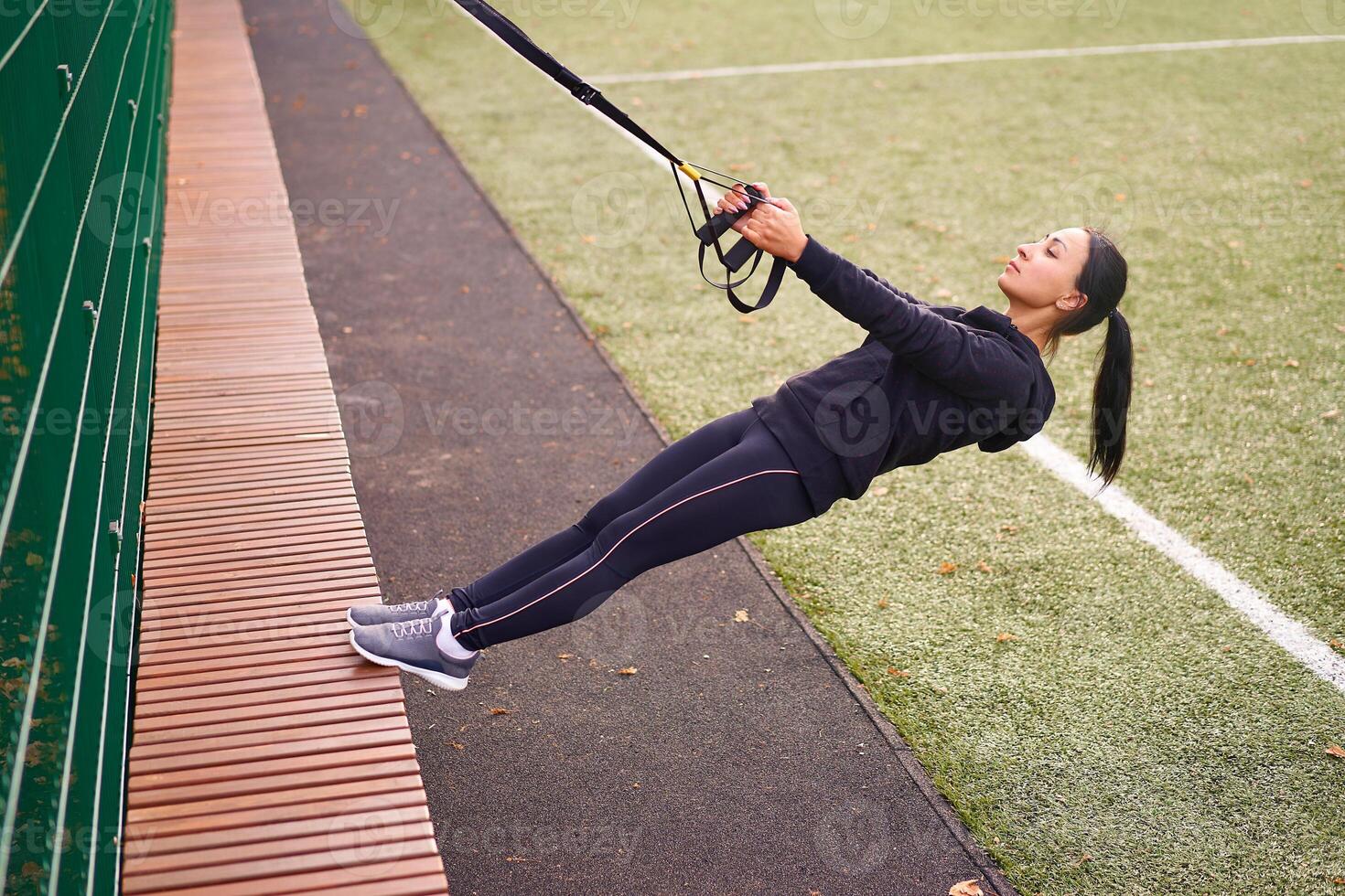 Girl athlete training using trx on sportground. Mixed race young adult woman do workout with suspension system. Healthy lifestyle. Stretching outdoors playground. photo