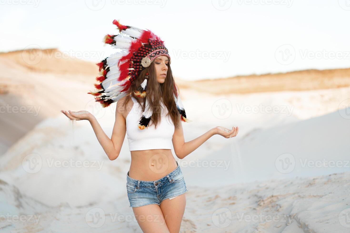 A beautiful young Caucasian girl in a white top and denim shorts. On her head  wearing an Indian hat Roach. Standing in the desert. photo