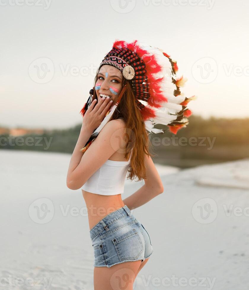 A beautiful young Caucasian girl in a white top and denim shorts. On her head  wearing an Indian hat Roach. Standing in the desert. photo