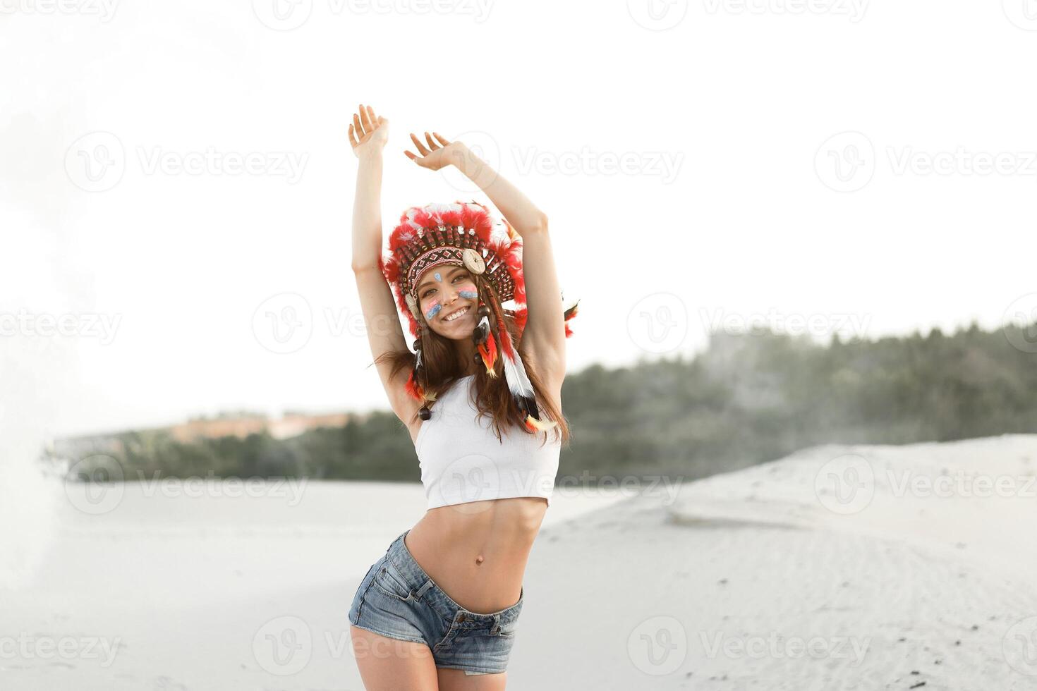 A beautiful young Caucasian girl in a white top and denim shorts. On her head  wearing an Indian hat Roach. Standing in the desert. photo