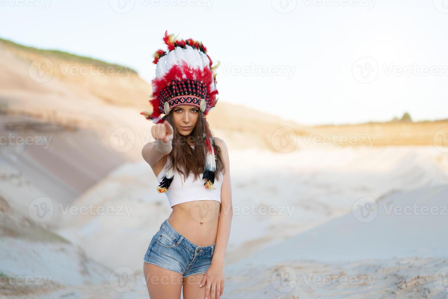 A beautiful young Caucasian girl in a white top and denim shorts. On her head  wearing an Indian hat Roach. Standing in the desert. photo