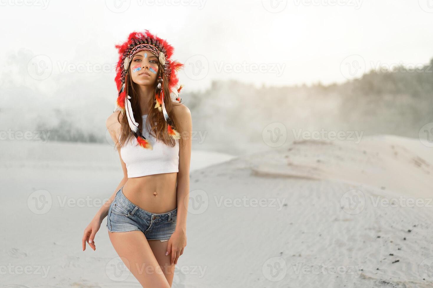 A beautiful young Caucasian girl in a white top and denim shorts. On her head  wearing an Indian hat Roach. Standing in the desert. photo