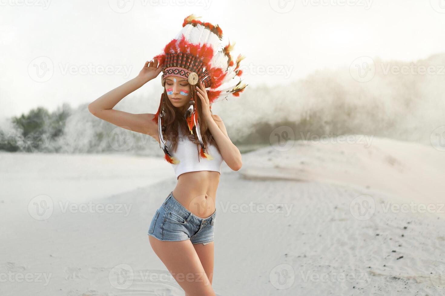 A beautiful young Caucasian girl in a white top and denim shorts. On her head  wearing an Indian hat Roach. Standing in the desert. photo