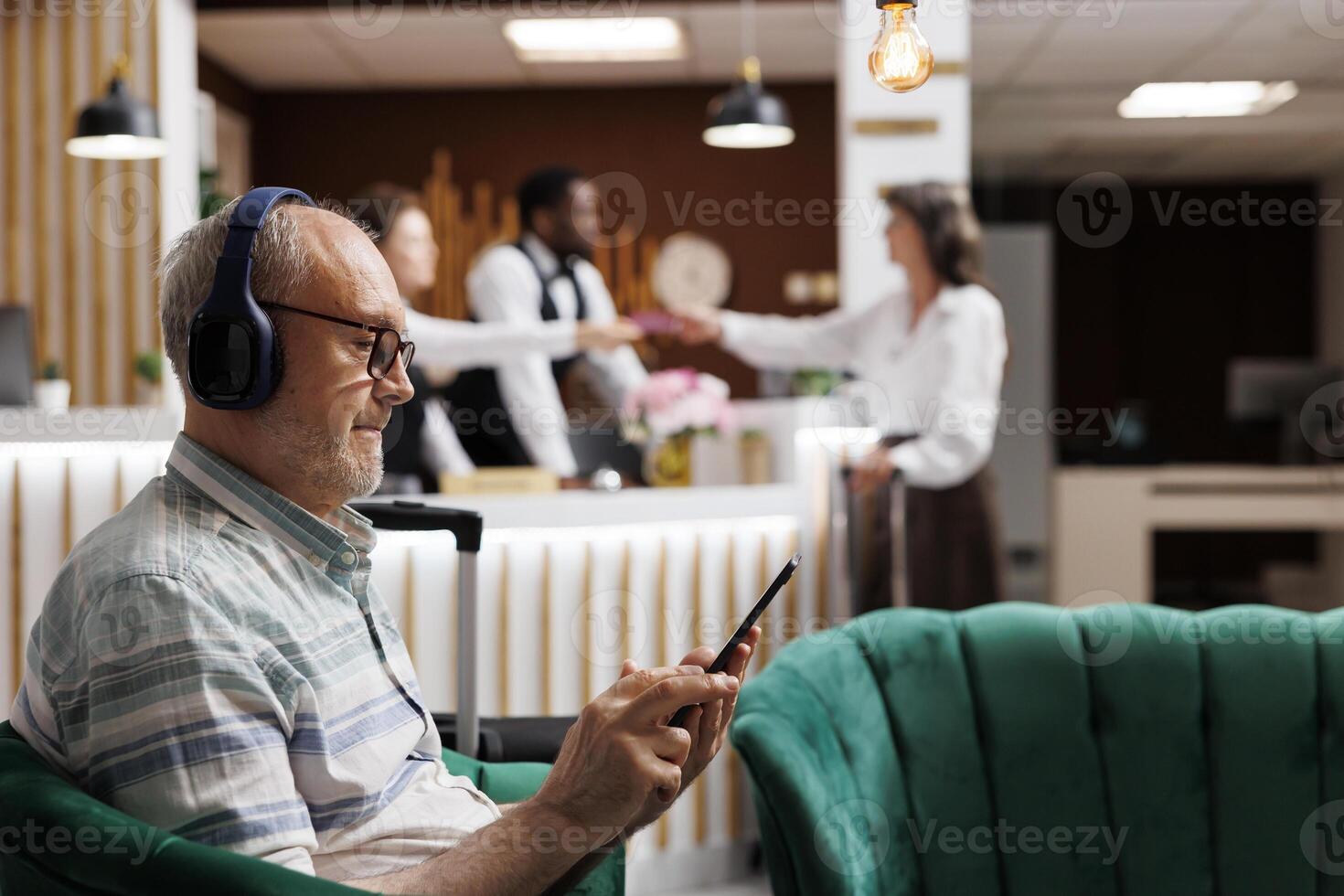 Detailed view of elderly male guest relaxing in cozy lounge area with tablet and wireless headset. Retired old man listening to music and surfing the net on digital device. Close-up, side-view. photo