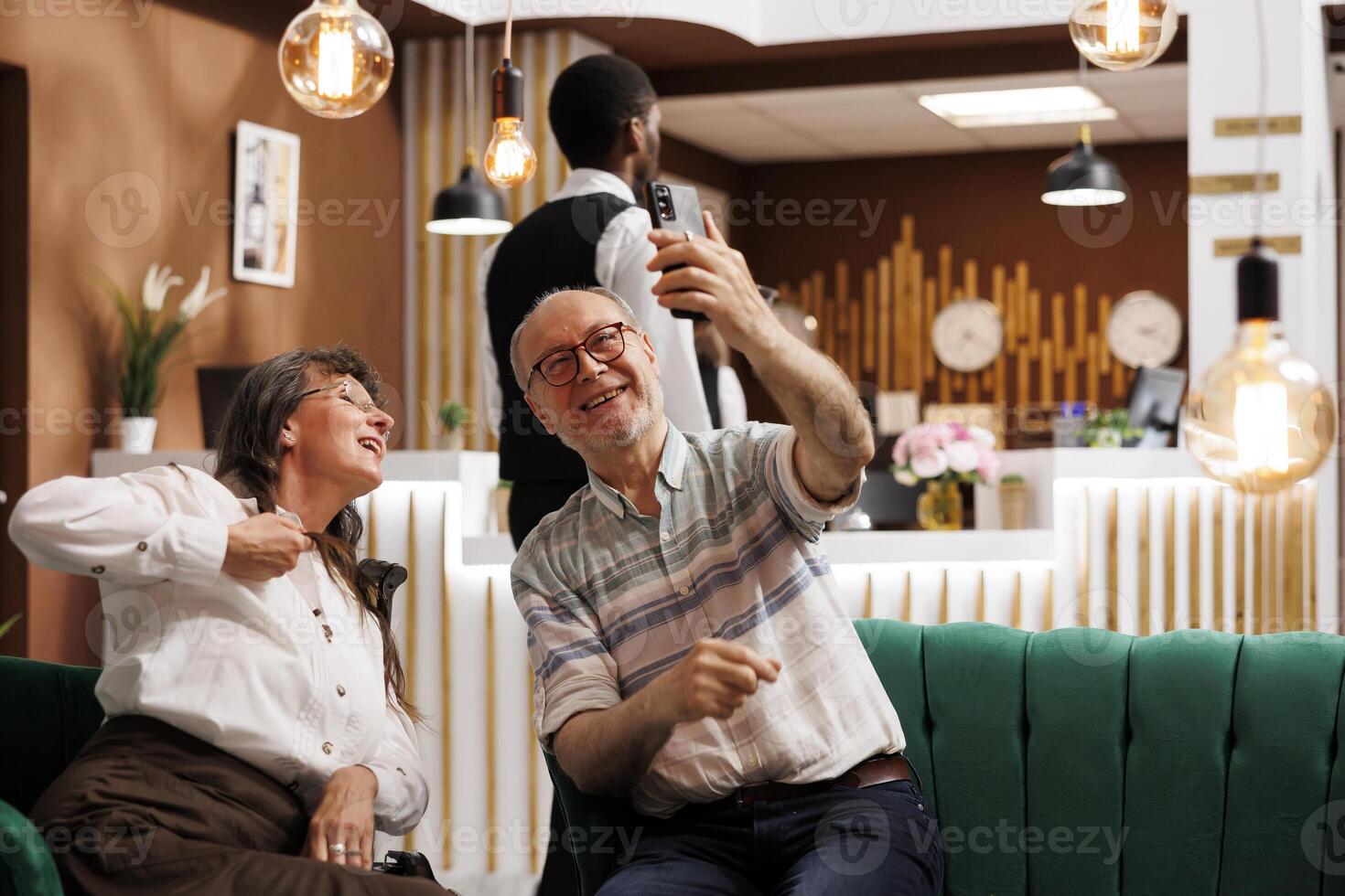 View from luxury hotel lobby of retired senior tourists making internet videocalls. Happy elderly couple on their honeymoon, connecting with family and friends through cell phone. photo