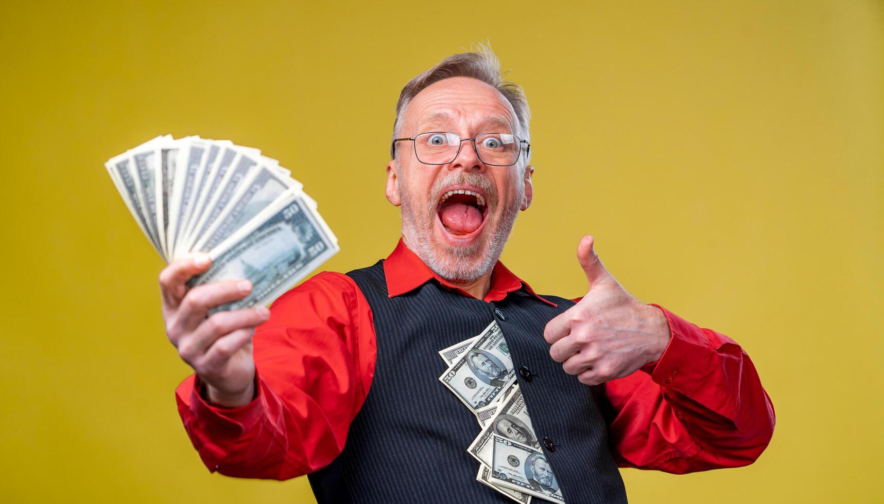 Old smiling grey-haired man in eyeglasses holding fan of dollars in front of the camera showing thumb up. Human emotions and facial expressions photo