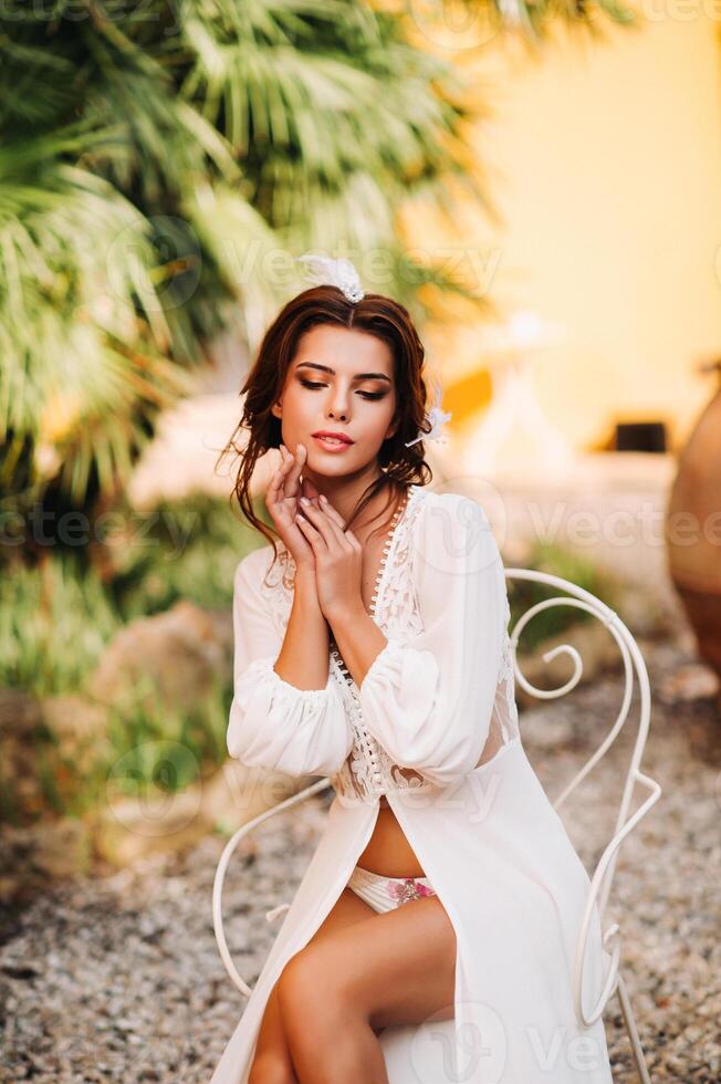 a beautiful bride is sitting on a chair and smiling in her underwear and dressing gown next to a Villa in Italy.morning of the bride in Tuscany photo