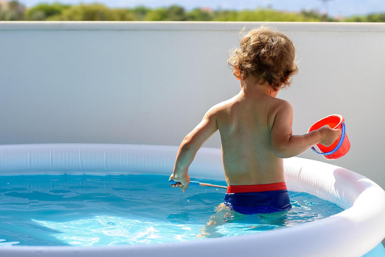 Back view of a little boy fishing with a net in the balcony pool water on a hot summer vacation day. He is holding a red bucket. View of crystal clean water surface, sun shine bright water-pool. photo