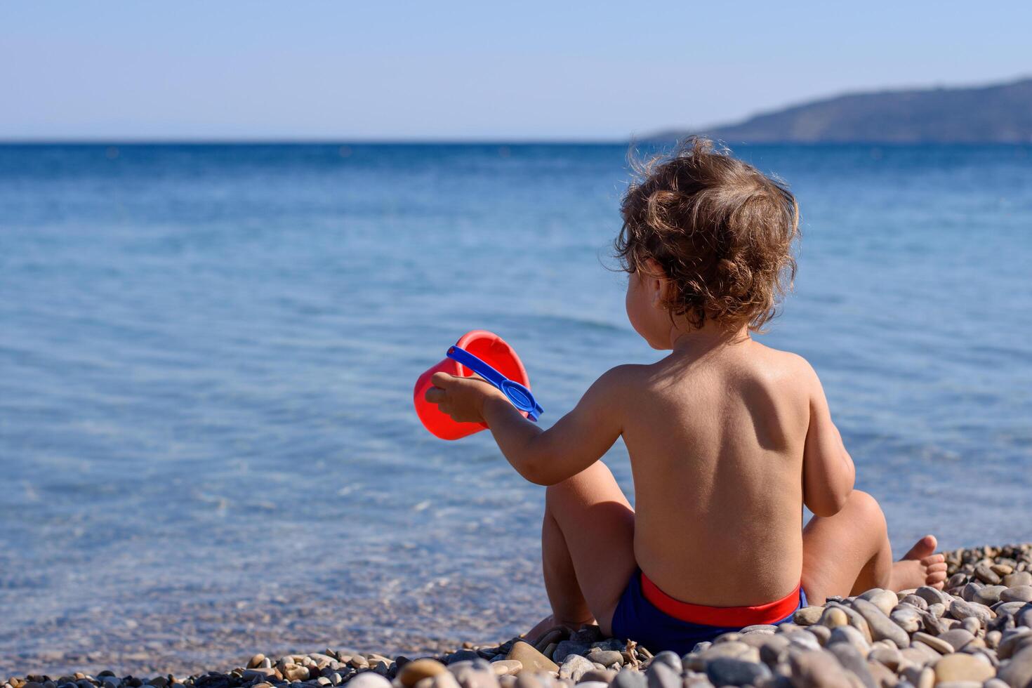 Back view of a little tan boy sitting on the seashore on a hot summer vacation day. He is holding a red bucket. View of crystal clean water surface Mediterranean, Agia Fotini beach Chios, Greece photo