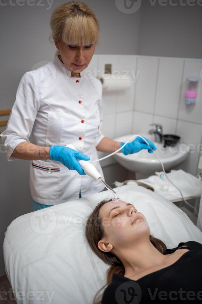 Beautician uses the darsonval device on the client's face. Professional skin care in the salon. The concept of cosmetology and rejuvenation. photo