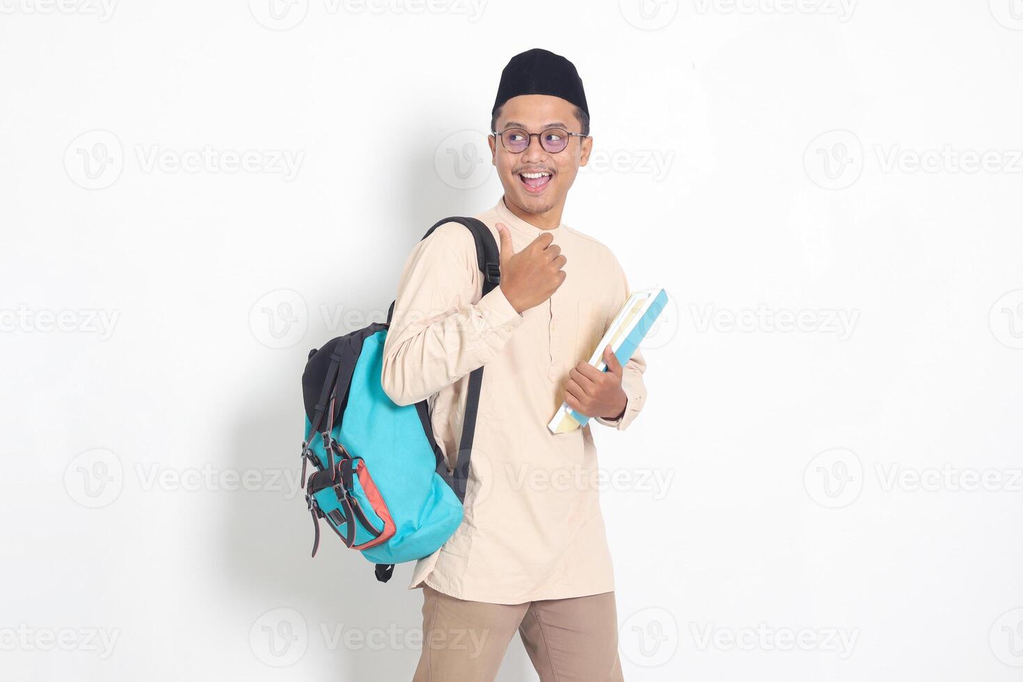 Portrait of excited student Asian muslim man in koko shirt with skullcap carrying backpack, holding school books, pointing to the side. Islamic education concept. Isolated image on white background photo