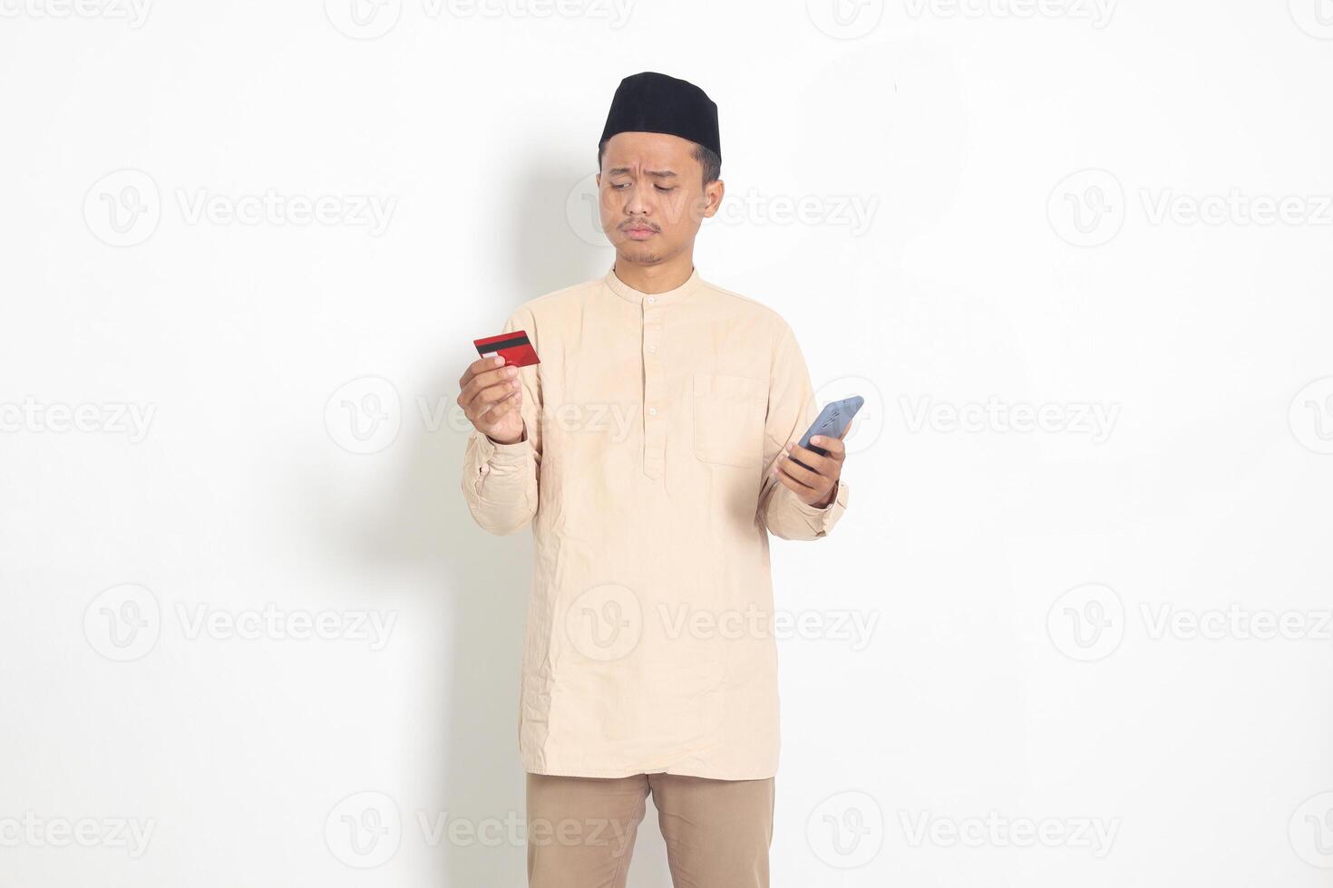 Portrait of upset Asian muslim man in koko shirt with skullcap holding a mobile phone and presenting credit card, deceived by online loans. Isolated image on white background photo