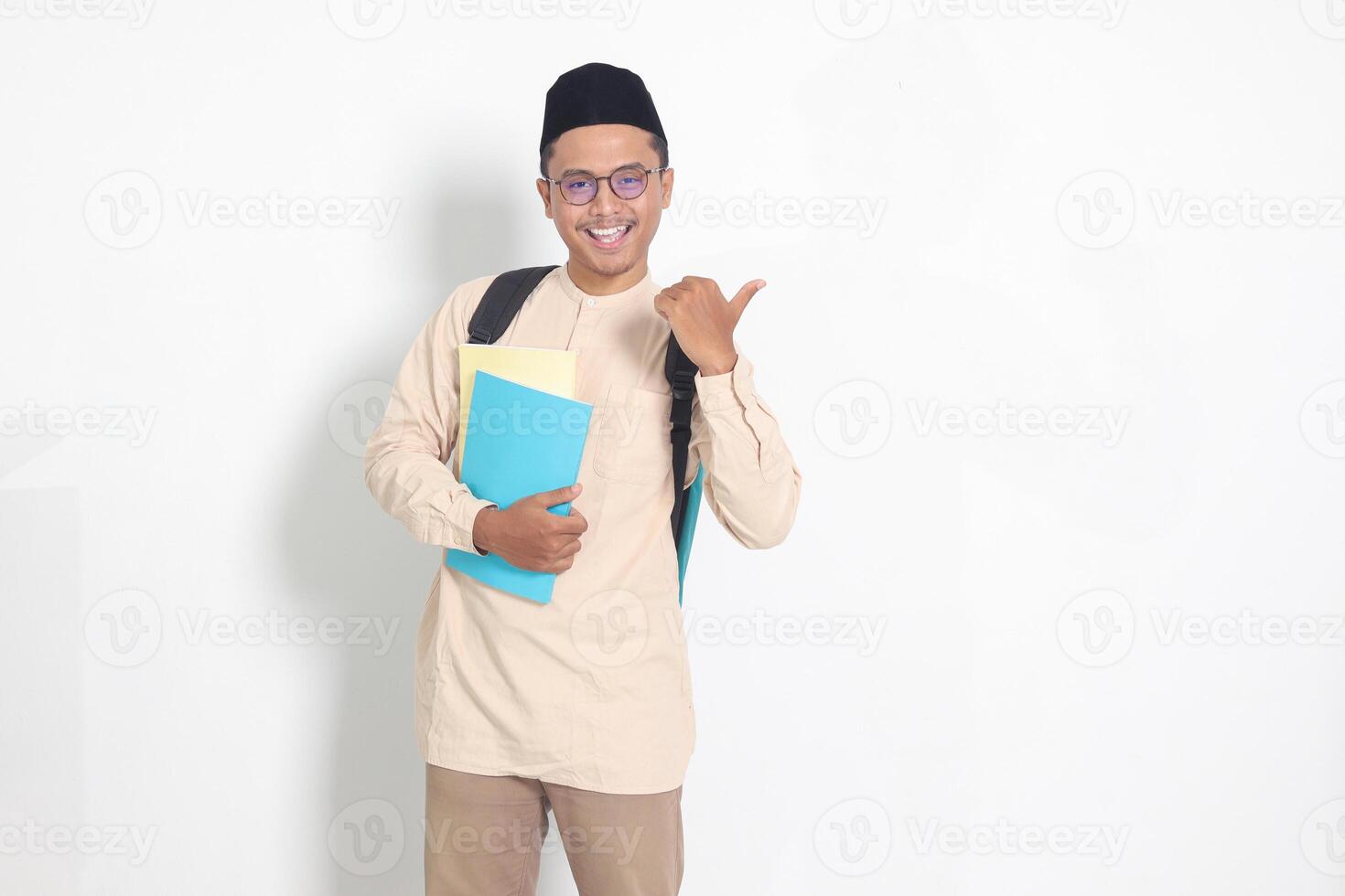 Portrait of excited student Asian muslim man in koko shirt with skullcap carrying backpack, holding school book, showing thumb up gesture. Islamic education concept. Isolated image on white background photo
