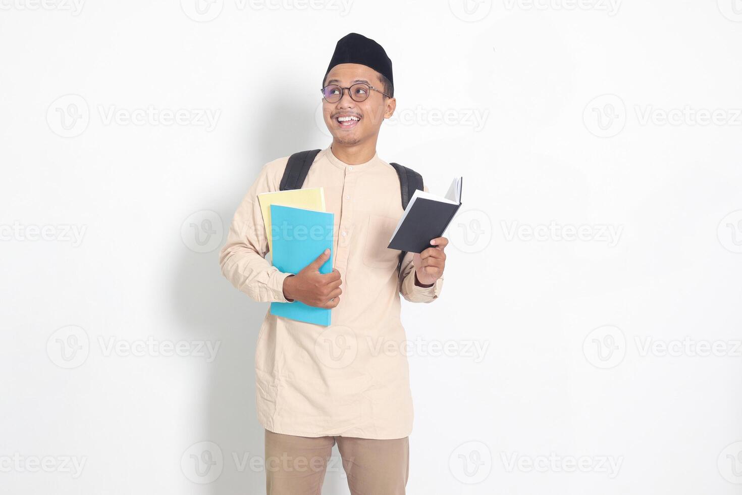 Portrait of excited student Asian muslim man in koko shirt with skullcap carrying school books, while reading his notebook. Isolated image on white background photo