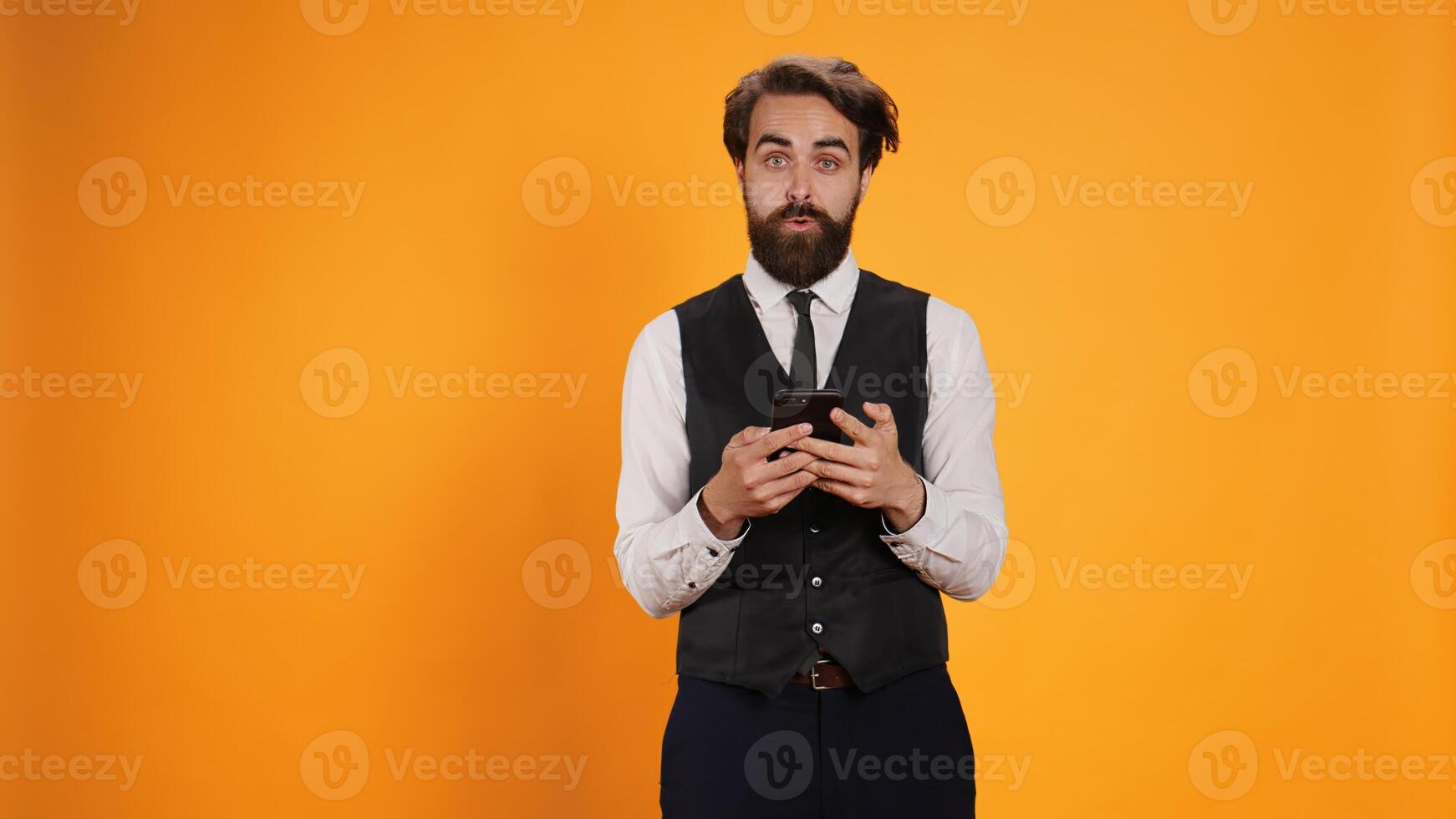 Diner employee messagging on smartphone, posing against yellow background. Experienced butler in formal clothes checking smartphone social media apps on work break, plate service. photo
