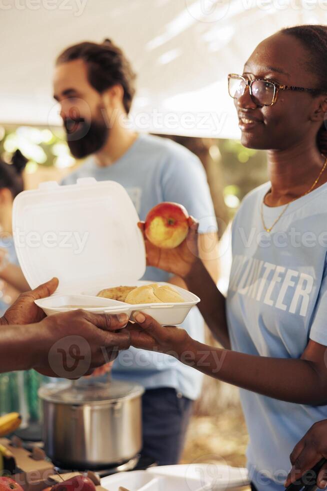 Close-up shot of generous black woman working in community food bank, serving free meals for the needy. Volunteering team of charity workers helping and sharing nutritrious items to homeless people. photo