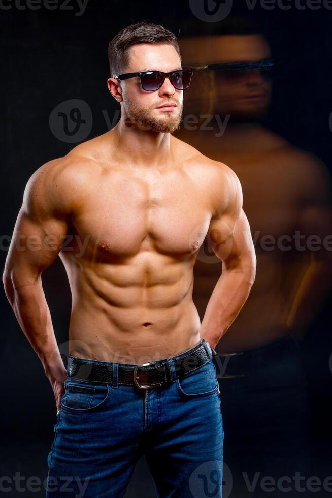 Beautiful young man with perfect body posing on dark background. Strong hands in back pockets. Guy in sunglasses. Dubling reflection. Studio photoshoot. photo