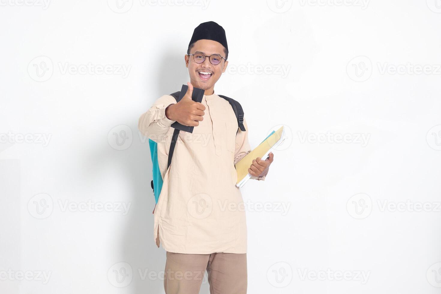 retrato de emocionado estudiante asiático musulmán hombre en koko camisa con casquete que lleva mochila, participación colegio libro, demostración pulgar arriba gesto. islámico educación concepto. aislado imagen en blanco antecedentes foto