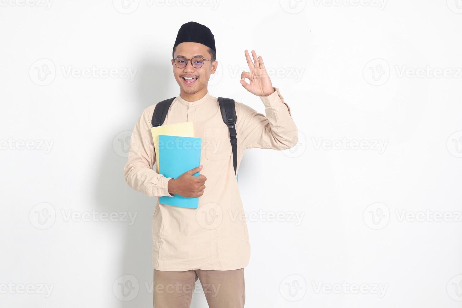 Portrait of excited student Asian muslim man in koko shirt with skullcap carrying backpack, holding school book, showing thumb up gesture. Islamic education concept. Isolated image on white background photo