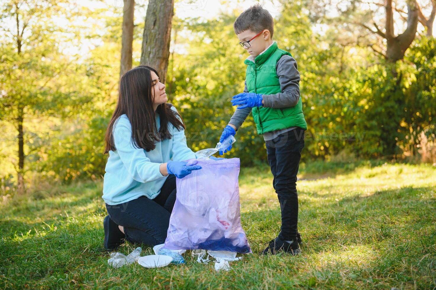 Woman volunteer and little boy picking up the plastic garbage and putting it in biodegradable trash-bag outdoors. Ecology, recycling and protection of nature concept. Environmental protection. photo