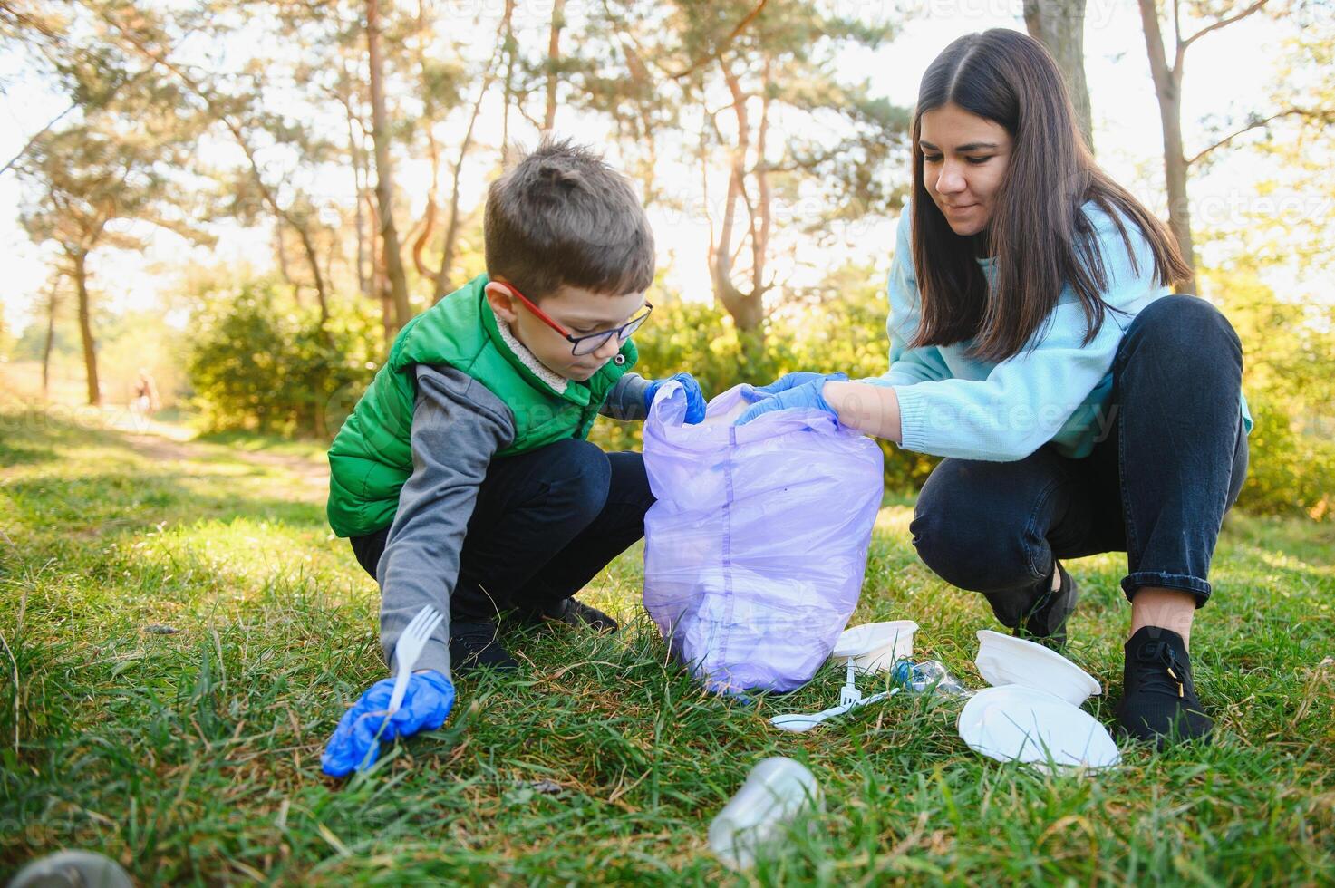 Woman volunteer and little boy picking up the plastic garbage and putting it in biodegradable trash-bag outdoors. Ecology, recycling and protection of nature concept. Environmental protection. photo