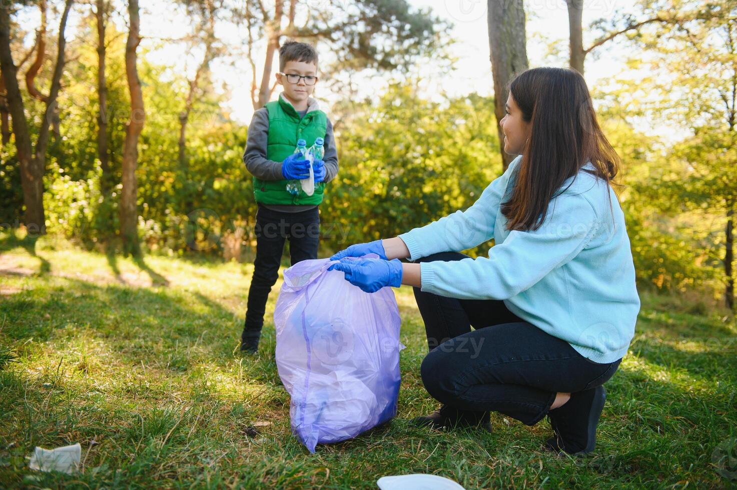 Woman volunteer and little boy picking up the plastic garbage and putting it in biodegradable trash-bag outdoors. Ecology, recycling and protection of nature concept. Environmental protection. photo