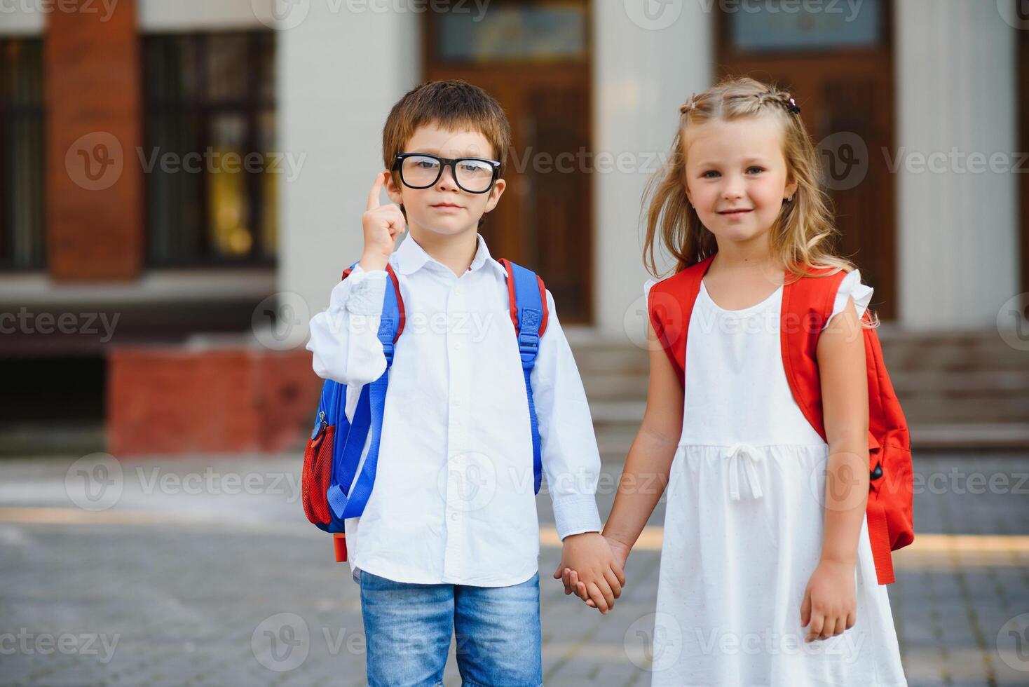 Two pupils of primary school. Boy and girl with school bags behind the back. Beginning of school lessons. Warm day of fall. Back to school. Little first graders. photo