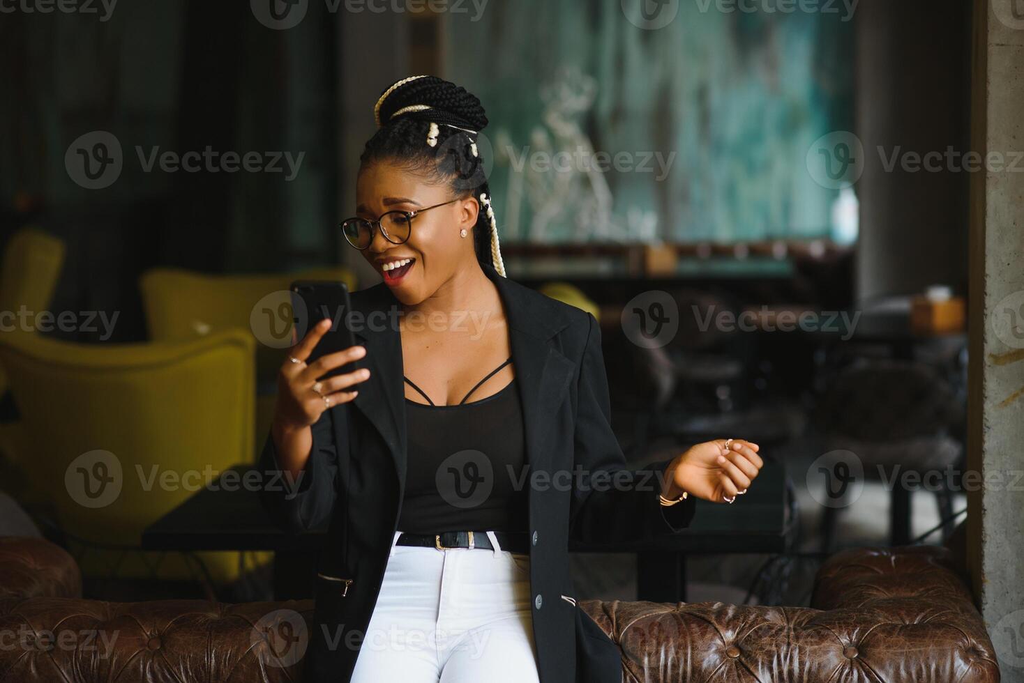 Cheerful dark skinned woman in trendy wear and spectacleson cafe posing for picture and media content for blog, smiling african american female 20s making selfie using mobile phone photo