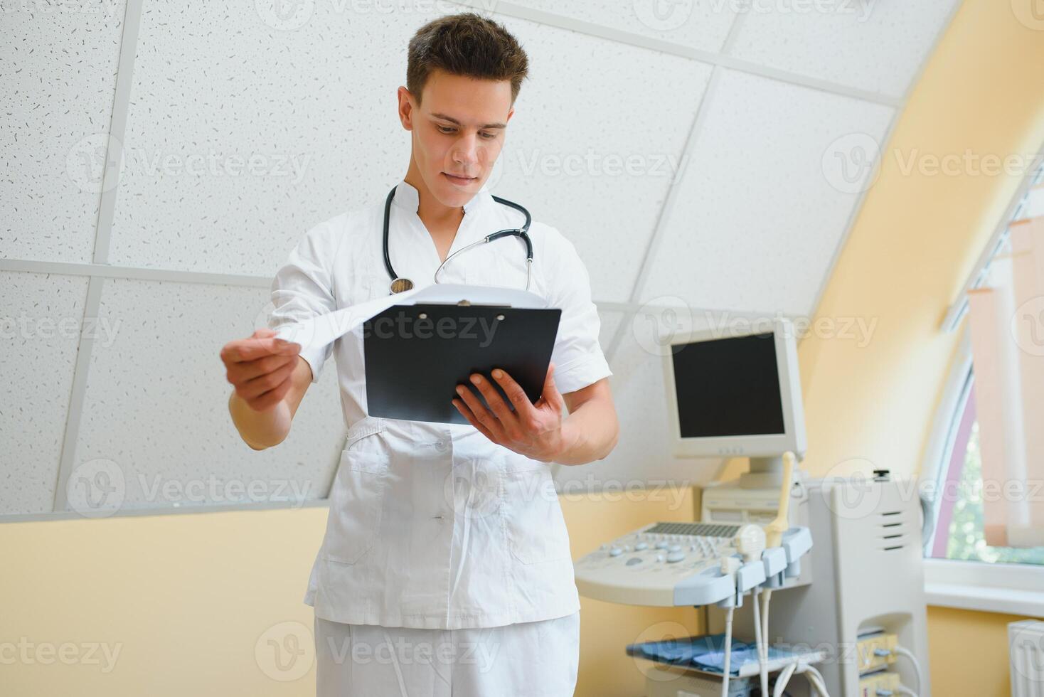 Portrait of a handsome young doctor next to an ultrasound scanning machine. He's squinting, looking at the camera with untrusting look, filling patient's history. photo