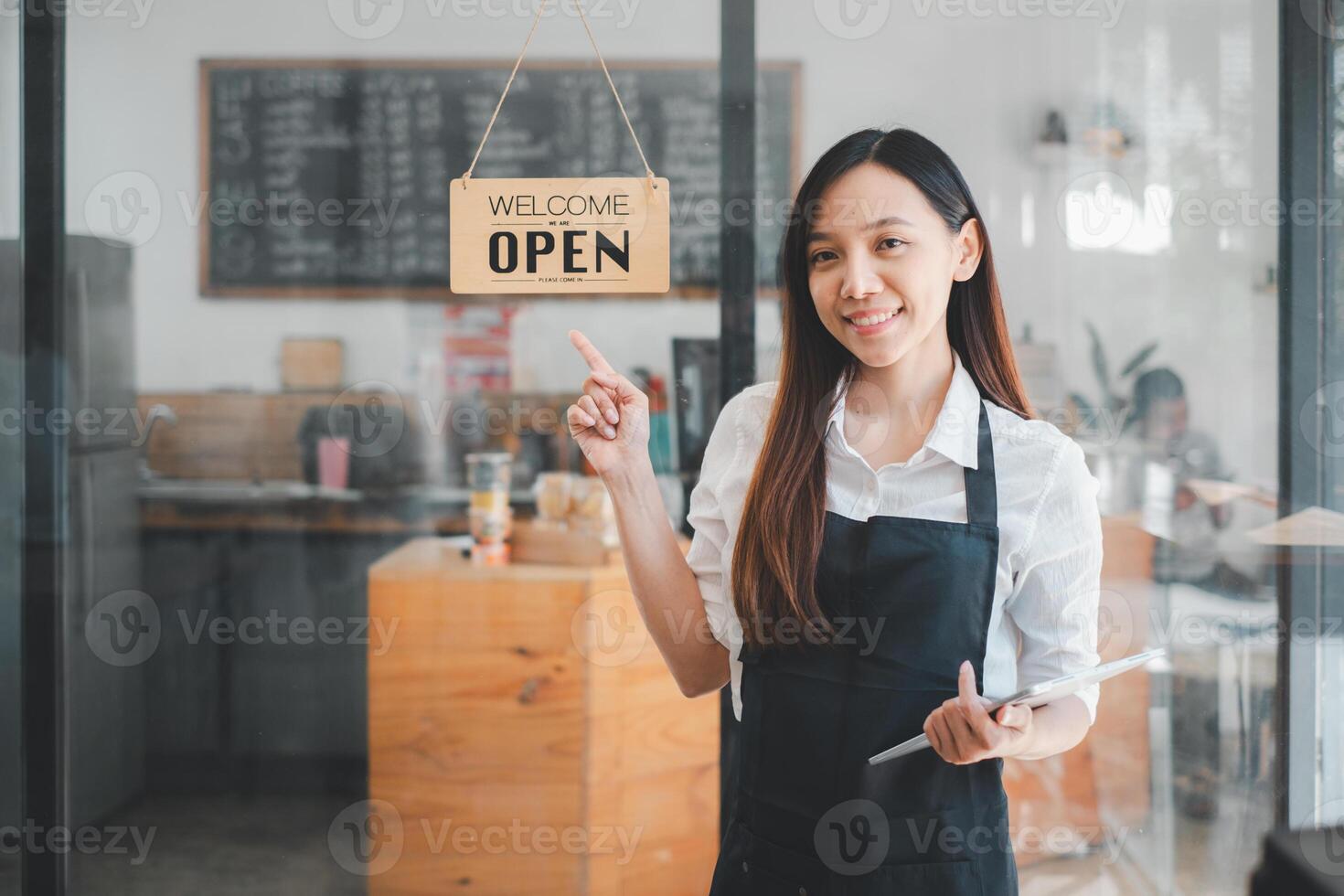 A cheerful cafe owner stands by the 'Welcome Open' sign at her storefront, tablet in hand, ready to greet customers into the warm atmosphere of her coffee shop. photo