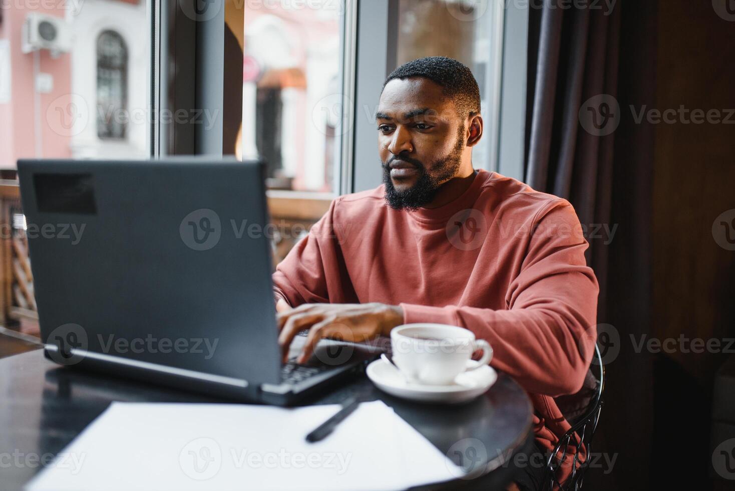 Portrait of thoughtful and concentrated young African American copy-writer, working on new articles using his laptop, and, looking at papers on a cafe table. photo