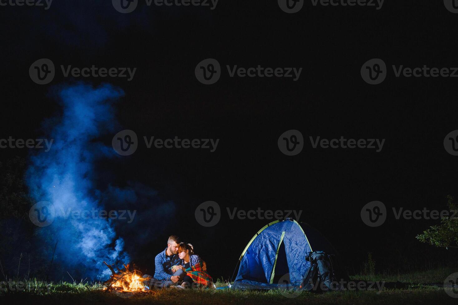 Young couple man and woman trevelers sitting near glowing tourist tent, burning campfire, on the top of mountain, enjoying beautiful view of night sky. photo