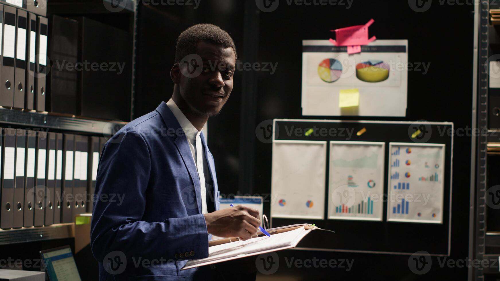 Determined black policeman standing and looking at camera in office full of evidence, files, and photos. Focused african american private investigator holding and reviewing crimanal records. Portait. photo