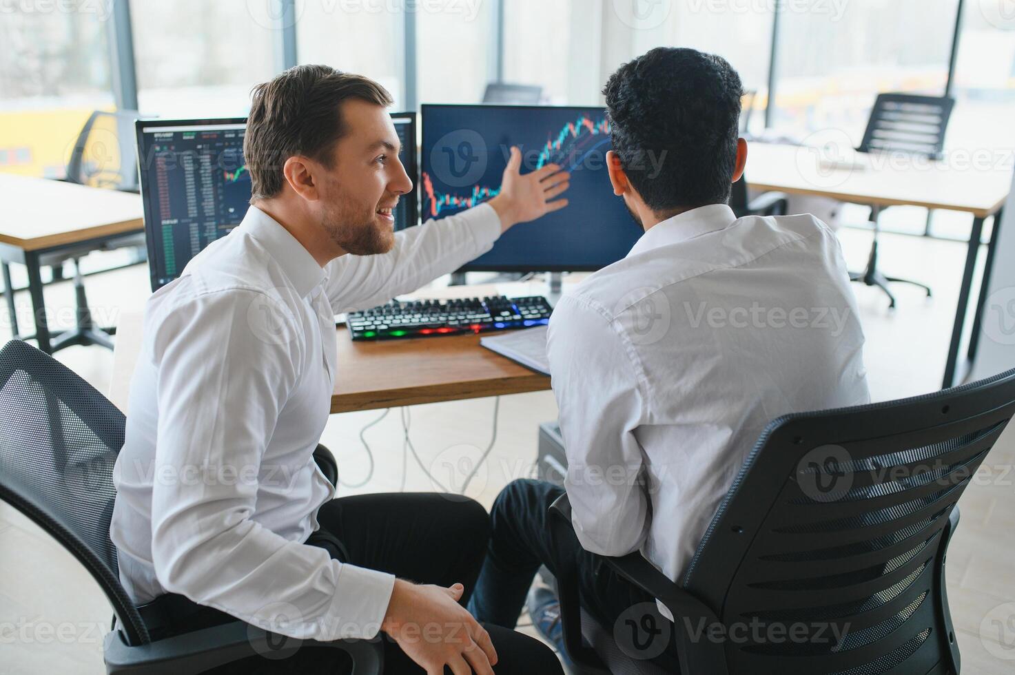 Two men traders sitting at desk at office together monitoring stocks data candle charts on screen analyzing price flow smiling cheerful having profit teamwork concept photo