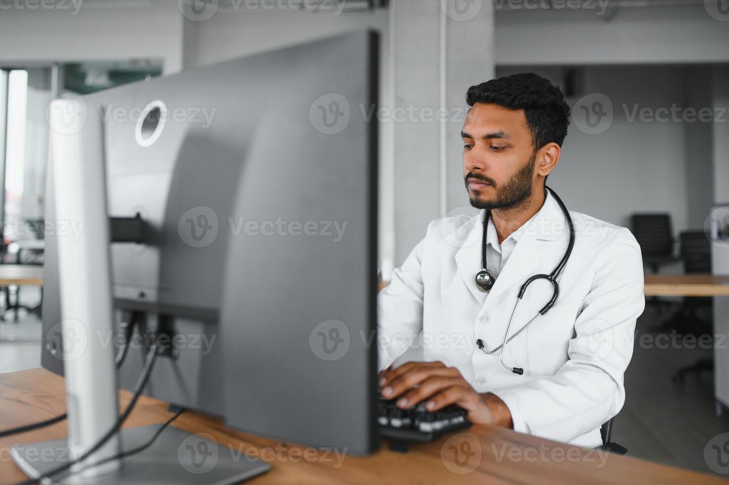 Indian male doctor physician wearing glasses, white medical gown and stethoscope sitting at the desk with the laptop in modern clinic and involved online video onference, consulting remotely. photo