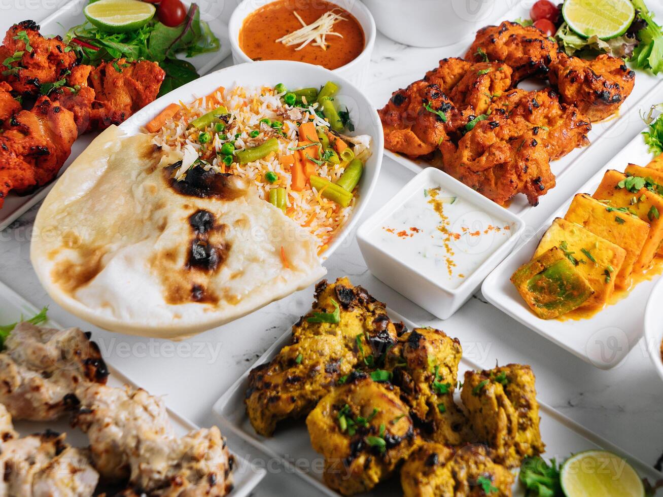 Assorted famous indian and pakistani food table vegetable biryani, Butter Chicken, paneer Chicken Tikka boti kebab, lime, Kali Mirchi, tomato sauce, raita, roti, salad, top view on grey background photo