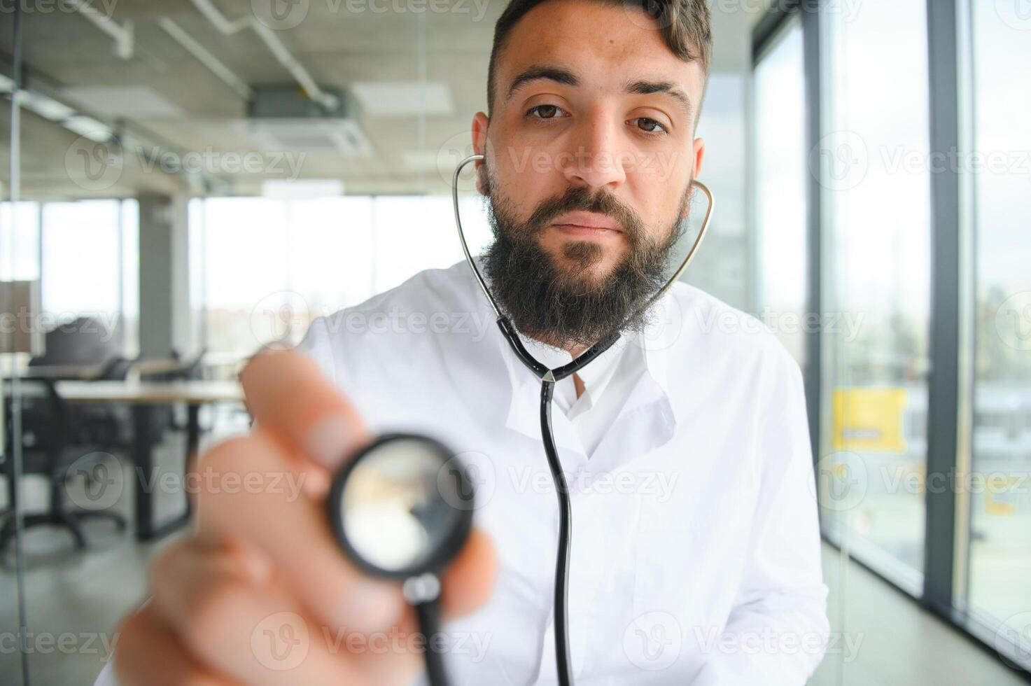 Young 30-aged handsome pleasant Arabic male doctor in white coat, posing at camera indoors. photo