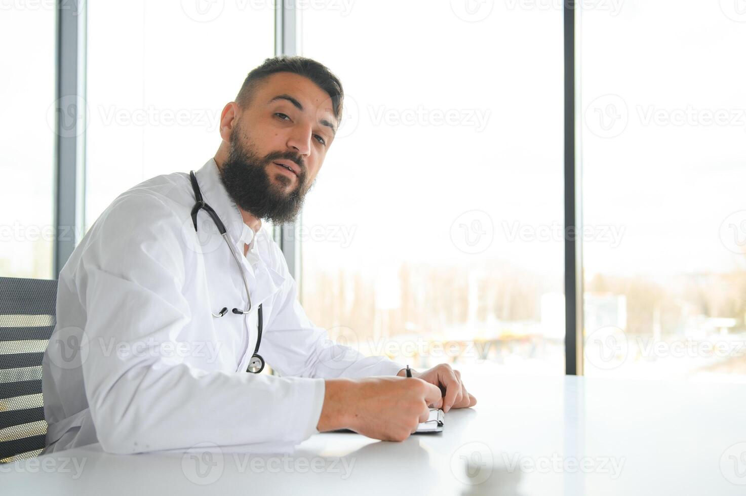 Young 30-aged handsome pleasant Arabic male doctor in white coat, posing at camera indoors. photo