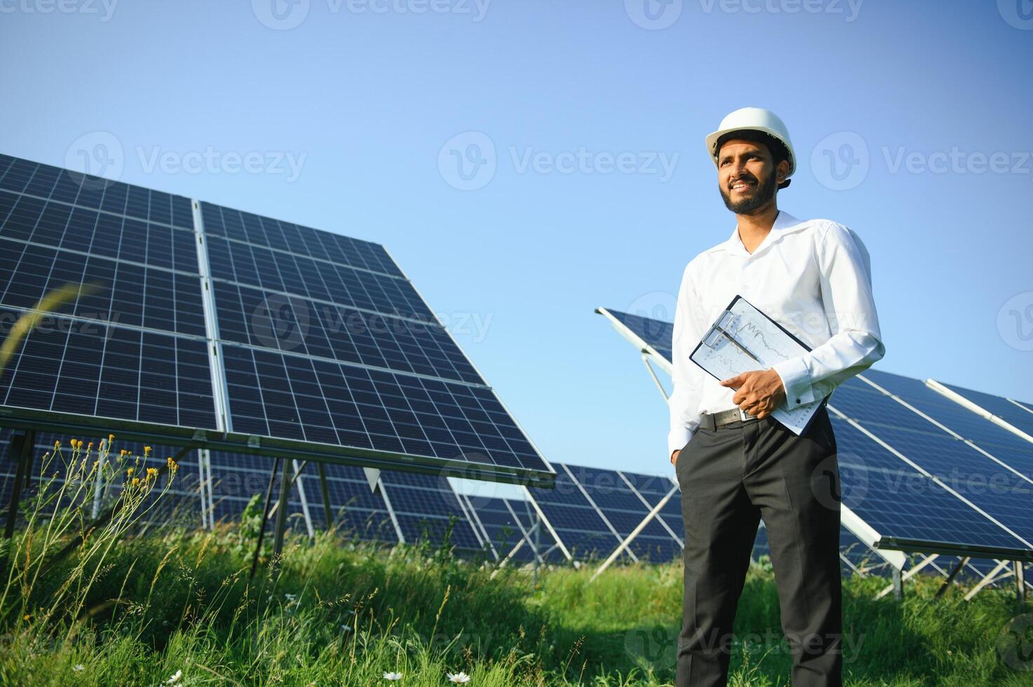 Portrait of Young indian man technician wearing white hard hat standing near solar panels against blue sky.Industrial worker solar system installation, renewable green energy generation concept. photo