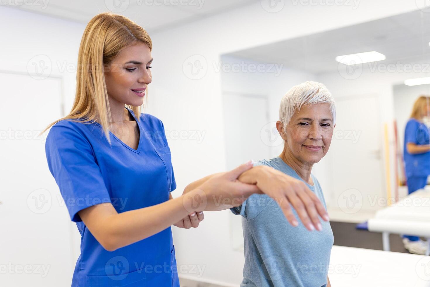 Caregiver helping contented senior woman exercise at clinic. Rehab for elder, senior medical care. Elder woman returning to good state of health photo