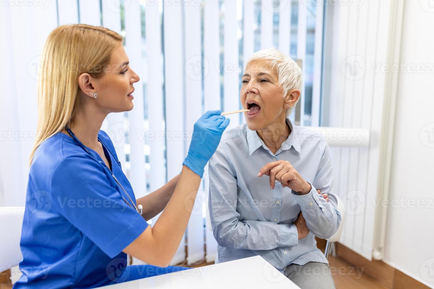 Senior patient opening her mouth for the doctor to look in her throat. Female doctor examining sore throat of patient in clinic. Otolaryngologist examines sore throat of patient. photo