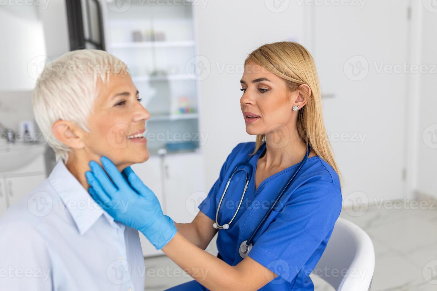 Friendly woman doctor wearing gloves checking sore throat or thyroid glands, touching neck of senior female patient visiting clinic office. Thyroid cancer prevention concept photo