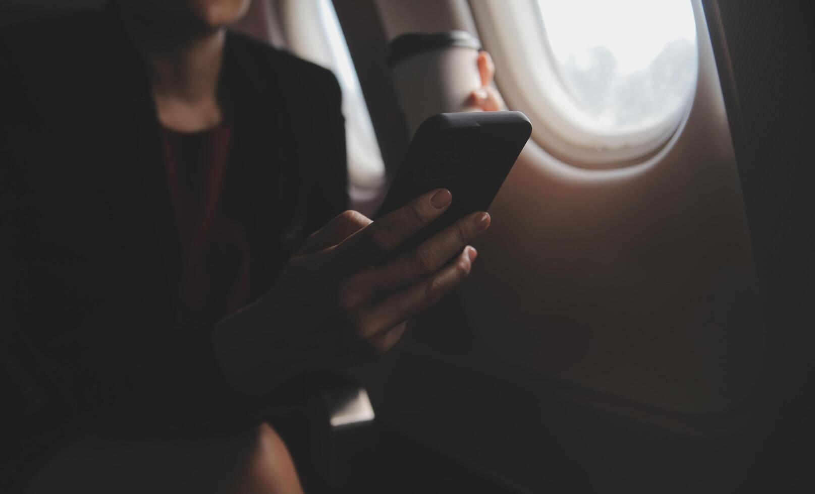 Blonde female tourist checking incoming notification on smartphone sitting on seat of airplane with netbook.Young businesswoman share media from telephone on laptop computer during plane flight photo