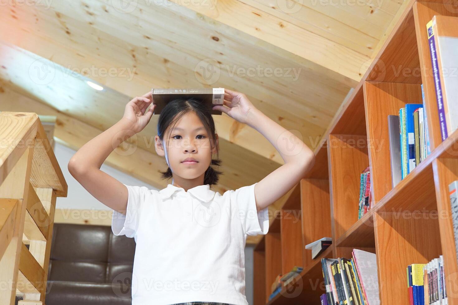 Asian girl takes a book from a bookshelf and puts it on her head. Concept, child research, education, development, brain, student, knowledge, learning. photo