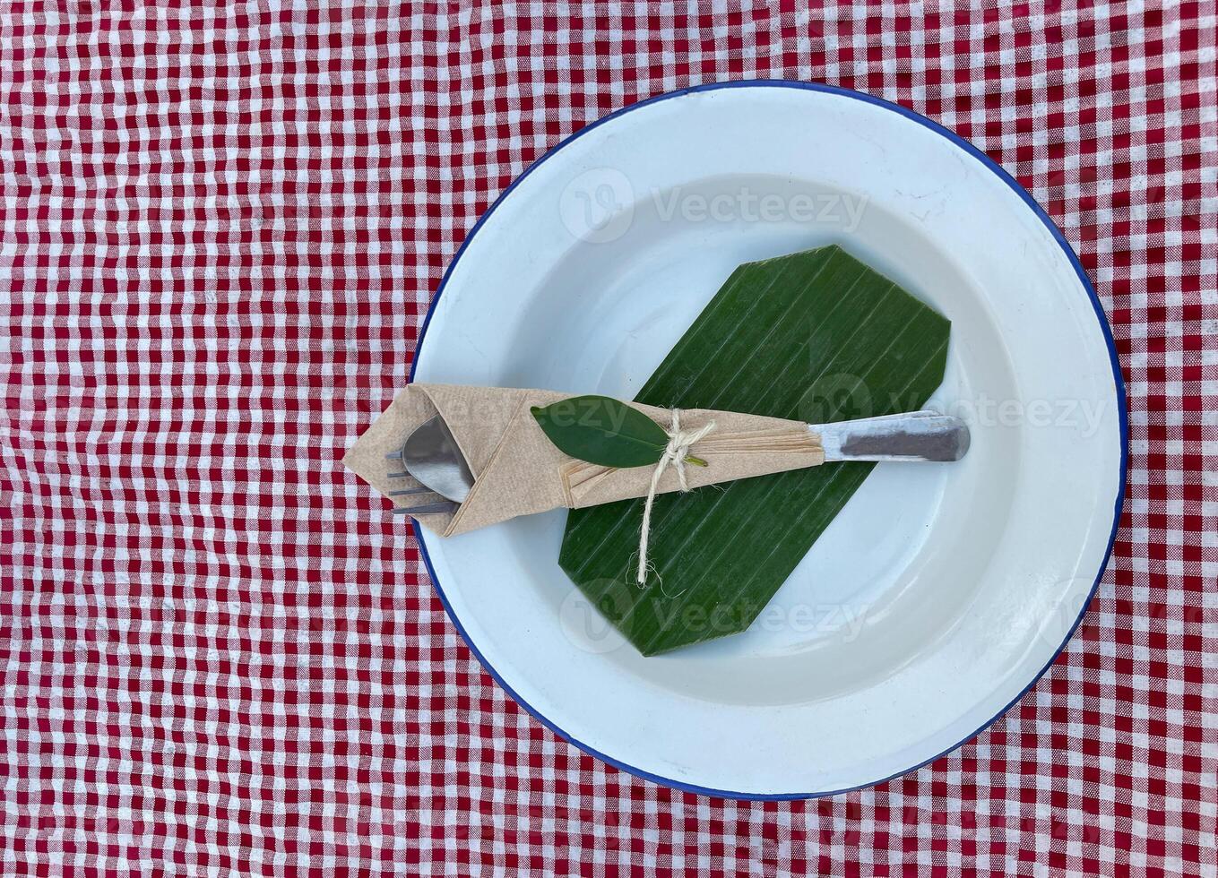 white galvanized plate is paired with a set of spoons and forks tied with hemp rope . Decorate the plate with banana leaves and place it on a red and white checkered tablecloth. photo