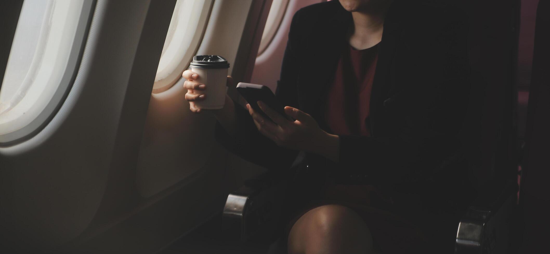 Blonde female tourist checking incoming notification on smartphone sitting on seat of airplane with netbook.Young businesswoman share media from telephone on laptop computer during plane flight photo