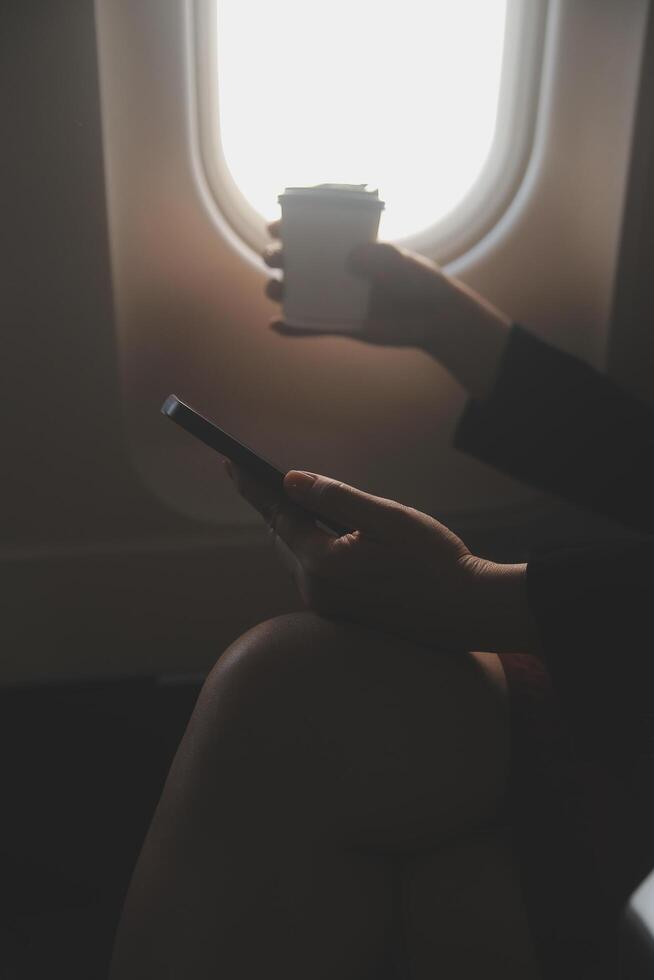 Blonde female tourist checking incoming notification on smartphone sitting on seat of airplane with netbook.Young businesswoman share media from telephone on laptop computer during plane flight photo