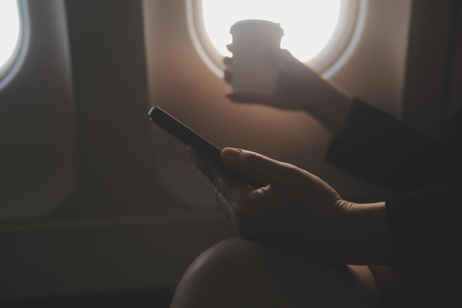 Blonde female tourist checking incoming notification on smartphone sitting on seat of airplane with netbook.Young businesswoman share media from telephone on laptop computer during plane flight photo