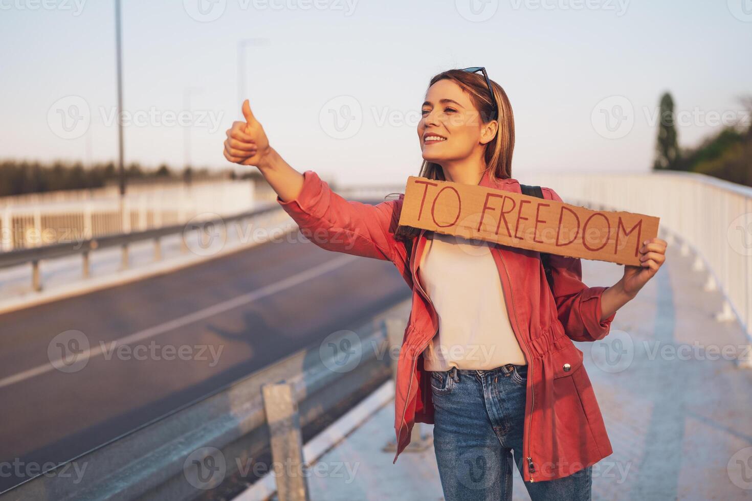 Woman is hitchhiking on roadside trying to stop car. She is holding cardboard with inscription. photo