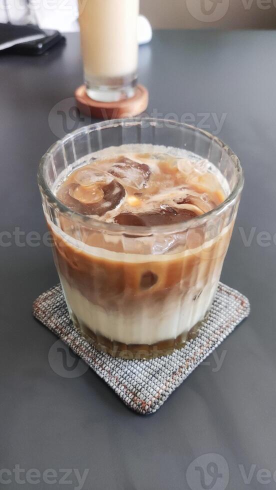 a glass of iced coffee on a coaster. The glass is tall and cylindrical with a straw in it. The coffee is light brown and there is a layer of ice at the bottom of the glass. photo